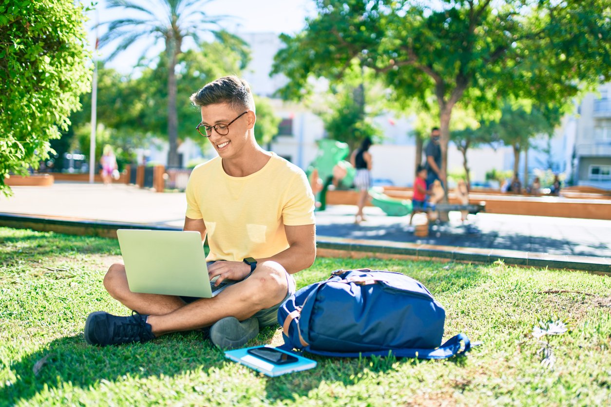 Smiling undergraduate student sitting in the grass on campus with his laptop